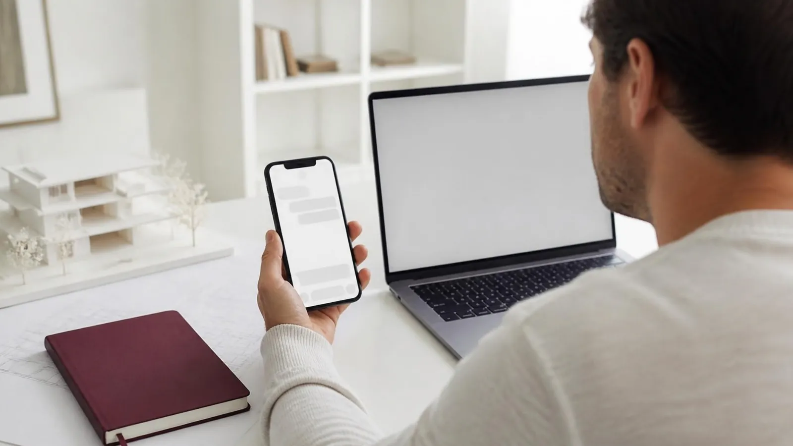 Architecte qui lit une demande de projet sur son téléphone, ordinateur portable et maquette blanche sur le bureau.